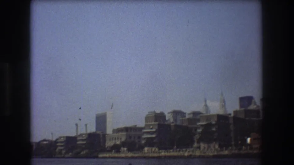 1976 New York City frame showing riverfront buildings, taller background towers, a hazy sky, and the dark edge of the 8mm contact-sheet frame.