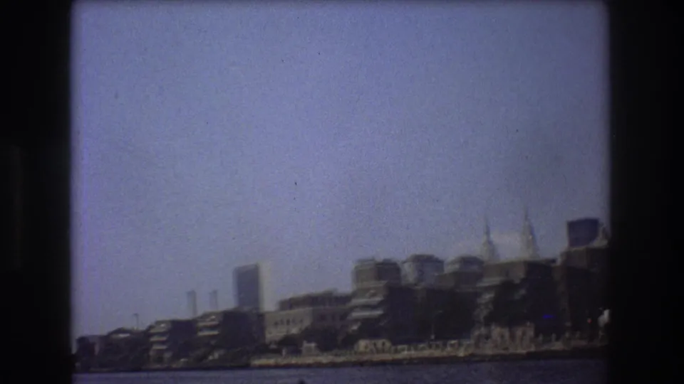 1976 New York City skyline seen from a boat across the water, with soft focus, haze, and dark 8mm frame edges.