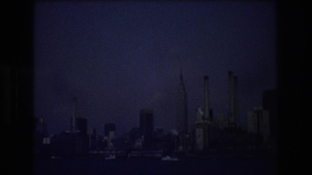 New York City waterfront skyline in 1976 viewed from the water, with industrial stacks and high-rises silhouetted in underexposed 8mm footage.