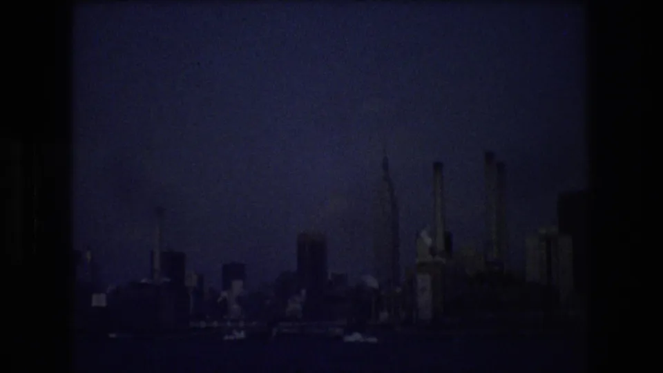 New York City waterfront skyline in 1976 viewed from the water, with industrial stacks and high-rises silhouetted in underexposed 8mm footage.
