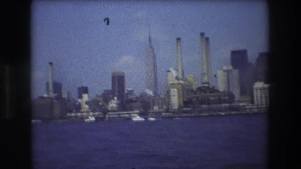 Boat-level 1976 New York City harbor view showing water, small boats, high-rises, and industrial smokestacks along the skyline.