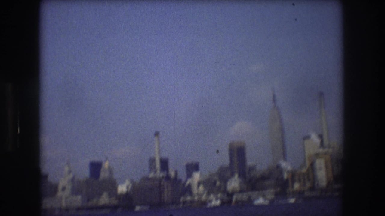 A hazy 1976 New York City skyline is seen from across the water, with distant towers, smokestack-like verticals, blue tint, and rounded 8mm frame edges.