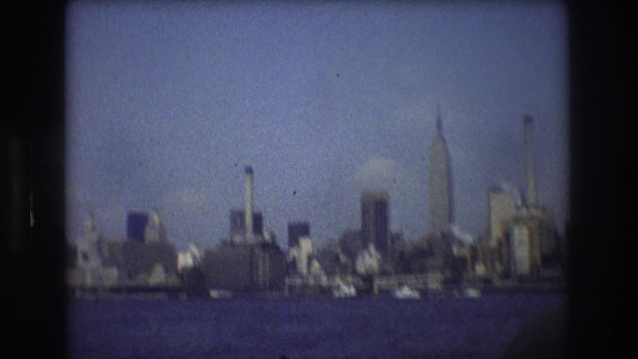 The New York City waterfront skyline appears in soft focus from a moving boat, with high-rise buildings, pale haze, blue water, and visible 8mm grain.