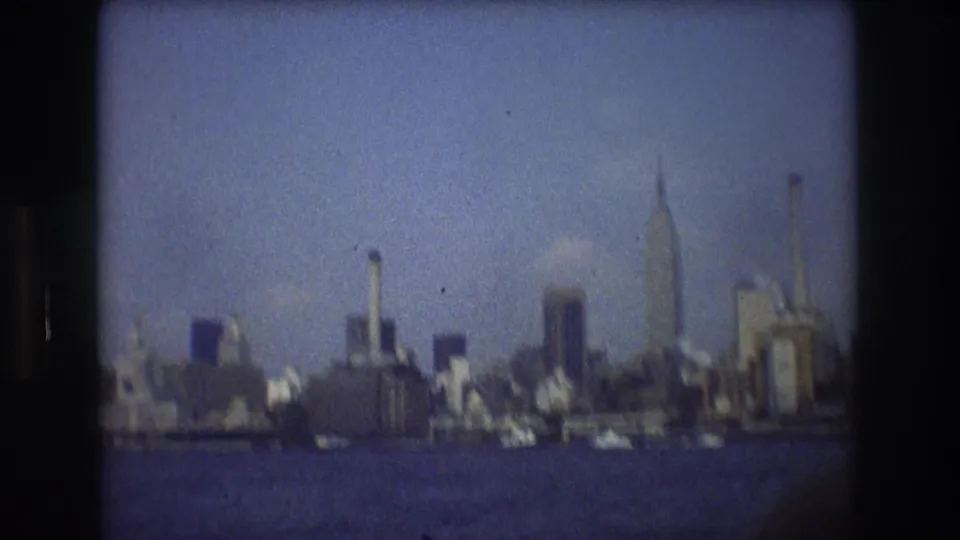 The New York City waterfront skyline appears in soft focus from a moving boat, with high-rise buildings, pale haze, blue water, and visible 8mm grain.