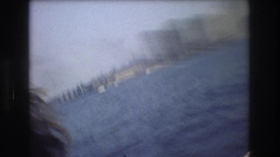 The camera tilts from a moving boat toward blue water and a blurred New York City shoreline, with the city forms softened by motion and haze.