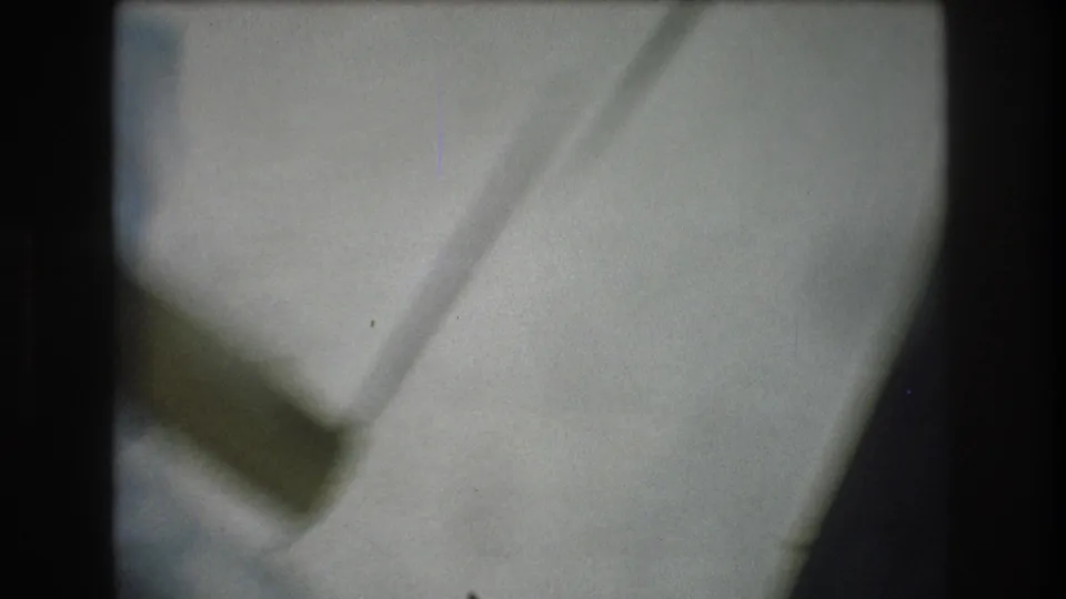 Out-of-focus 1976 8mm frame with a pale indoor plane, a diagonal line or edge, and a dark vertical area from the moving camera view.