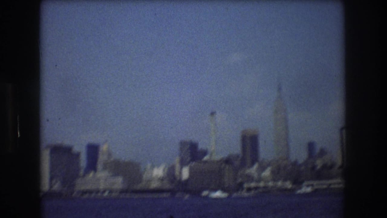 New York City skyline in 1976 viewed from across the water, showing tall buildings and a hazy blue sky in soft 8mm color.