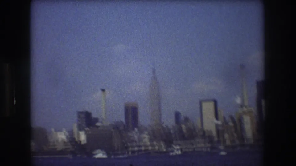 Soft 8mm frame of the New York City skyline, with harbor water, small bright boats, and several tall buildings visible through haze.