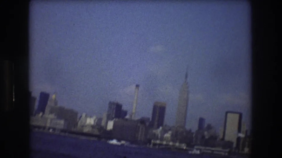 Handheld 1976 8mm view of the New York City skyline across dark blue harbor water, slightly tilted with visible grain and frame borders.