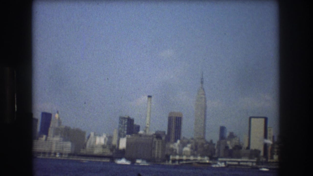 1976 8mm view of the New York City skyline from the water, with the Empire State Building rising above mid-century buildings under a blue sky.