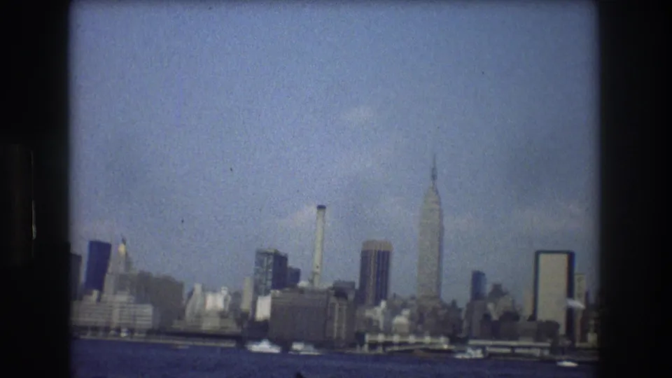1976 8mm view of the New York City skyline from the water, with the Empire State Building rising above mid-century buildings under a blue sky.