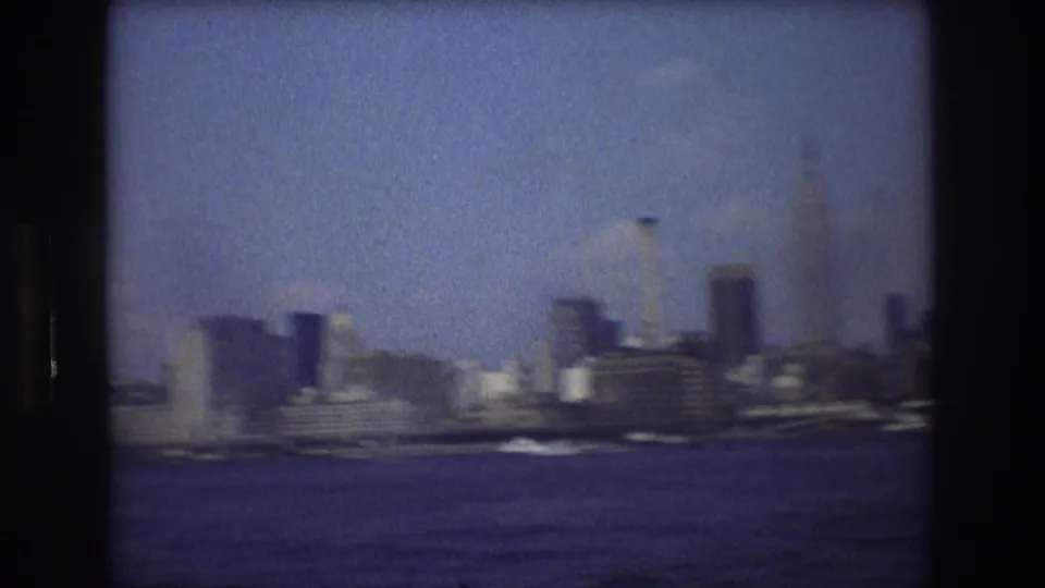 Motion-softened 1976 home-movie frame showing New York City waterfront buildings, small boats, and a distant skyline under a pale blue sky.