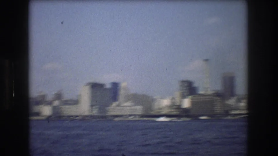 Late-frame 8mm view from the water toward New York City waterfront buildings, with choppy water, soft focus, and skyline forms under blue sky.