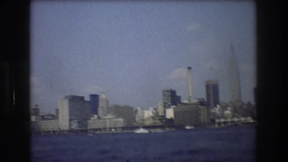 1976 8mm home-movie frame showing a hazy New York City skyline across open water, with high-rise buildings softened by grain and focus.
