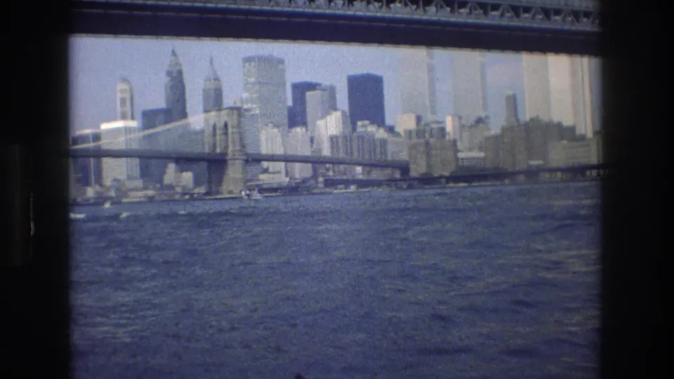 1976 8mm frame of a large New York City suspension bridge over water, with a dense Manhattan skyline visible through haze in the background.