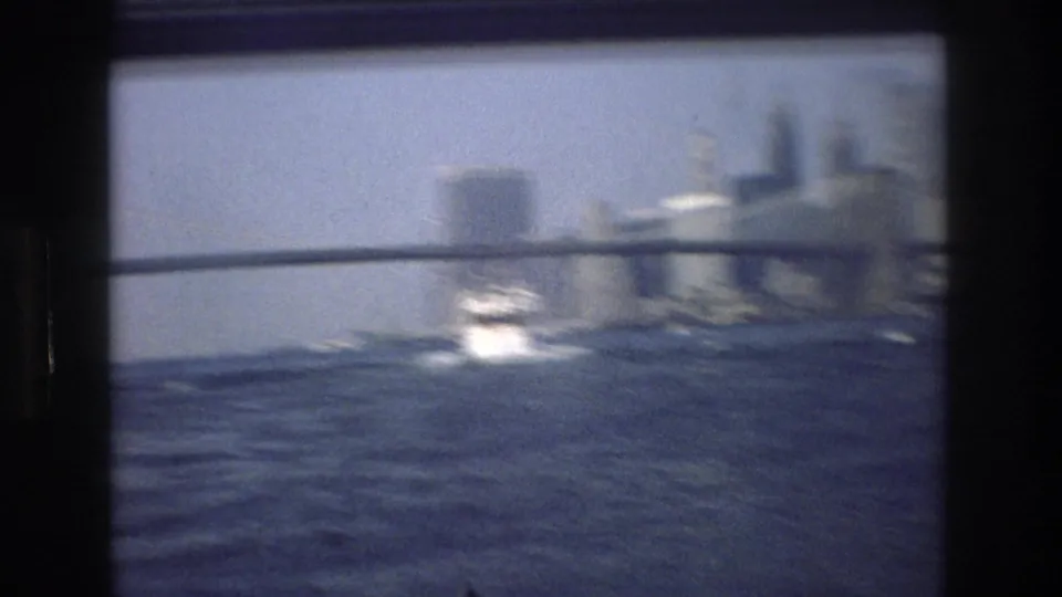 Soft, motion-blurred 1976 New York City home-movie frame showing a small white boat on the water, with a bridge and skyline faintly behind it.