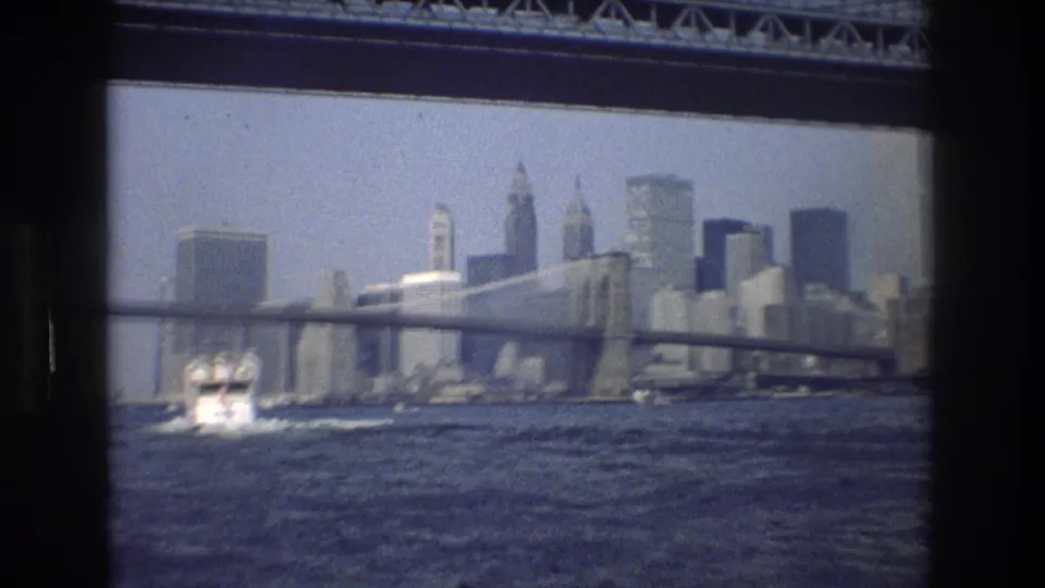 1976 New York City 8mm frame showing the Manhattan skyline beyond a bridge and harbor water, with a small boat wake in the foreground.