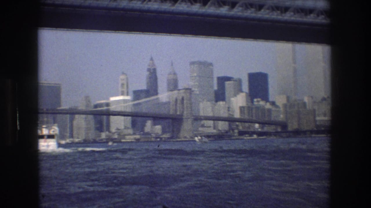 New York City skyline seen across water beneath a suspension bridge.