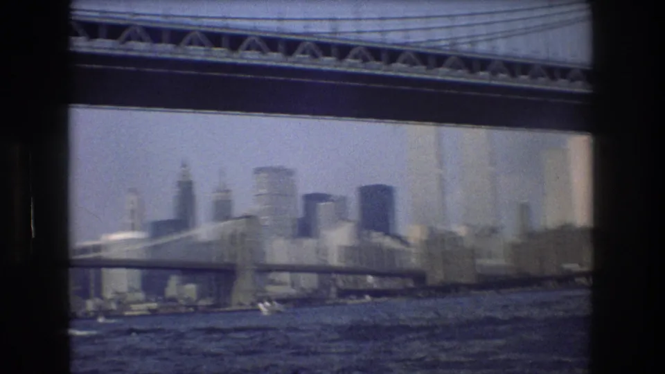 A soft 8mm view of New York City from the water, with a large suspension bridge in front of the skyline.