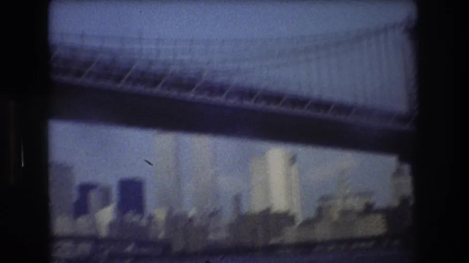 The camera passes close under a suspension bridge, partially obscuring the New York City skyline in a blurred 8mm frame.
