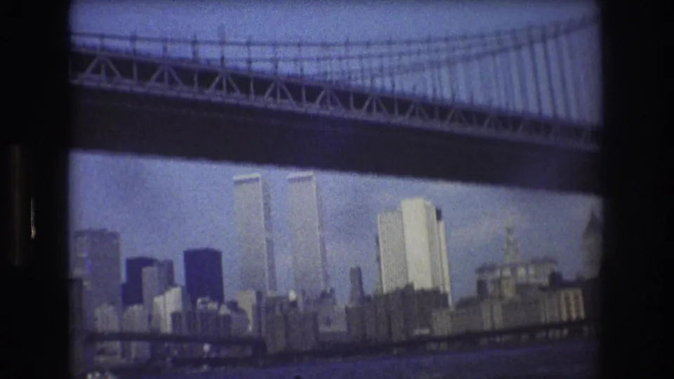 1976 8mm view of New York City's Manhattan skyline, with the Twin Towers visible below the dark underside of a large suspension bridge.