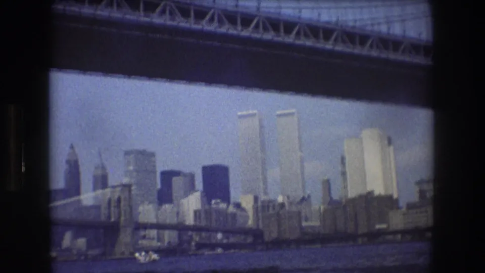 1976 New York City harbor view showing the Manhattan skyline, Twin Towers, bridge structure, waterfront buildings, and a small boat crossing the foreground.