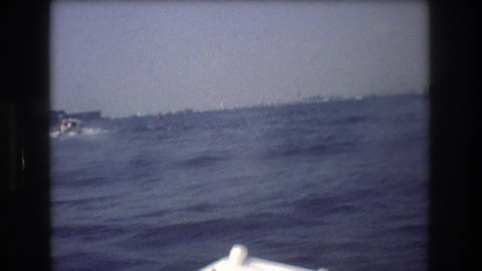 1976 8mm harbor shot in New York City, looking across open water toward a distant shoreline with a small boat near the left side of the frame.