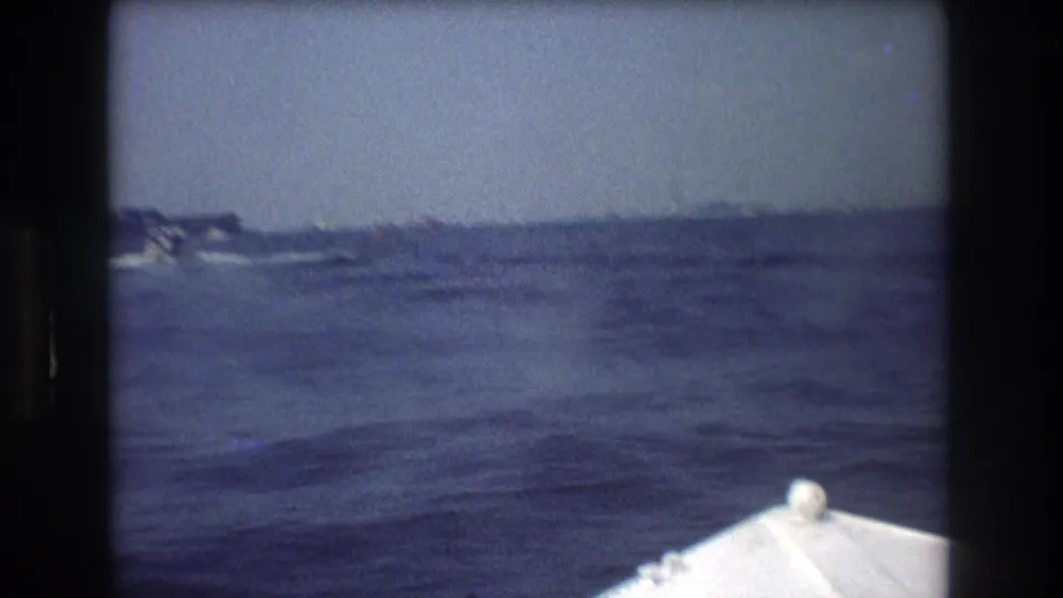1976 8mm frame showing a passenger view from a boat, with the white bow in the foreground and distant vessels or shoreline shapes across choppy water.