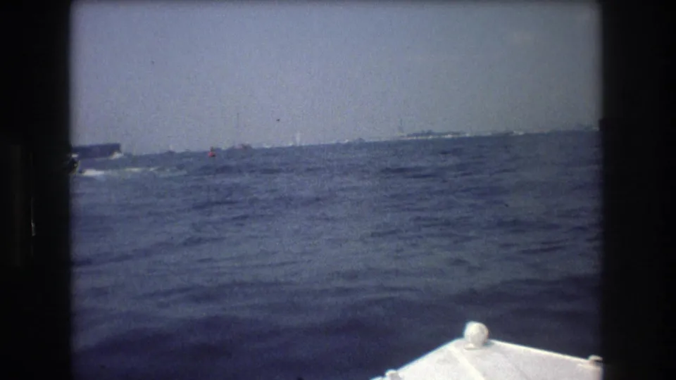 1976 8mm home-movie frame from a moving boat, with a partial white bow in the lower frame and rough water leading toward a faint distant vessel or shoreline.