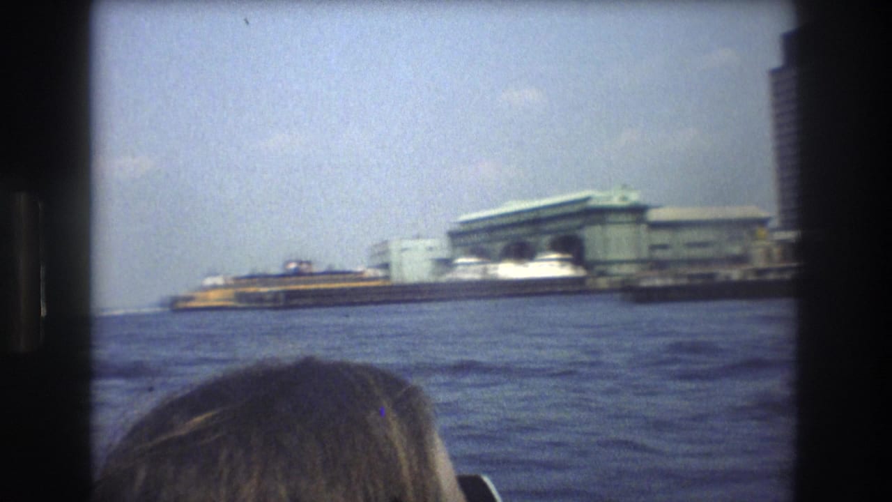 1976 New York City harbor view from a moving boat, showing choppy water, a waterfront terminal, tall buildings, and the top of a passenger's head at the lower edge.