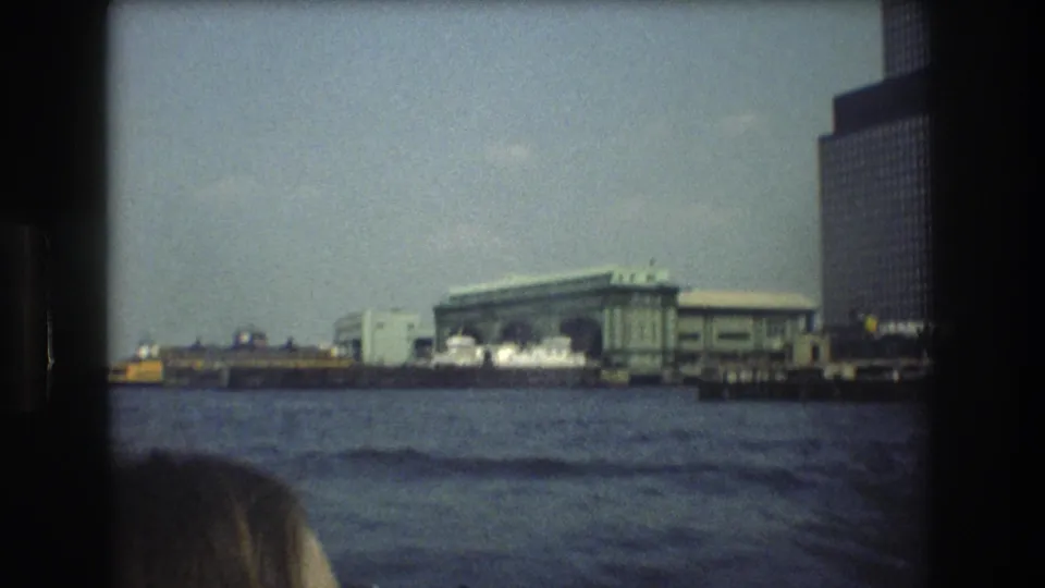 New York City harbor scene from the supplied 1976 footage, with a terminal across the water, modern high-rise buildings, and a partial passenger head in the foreground.