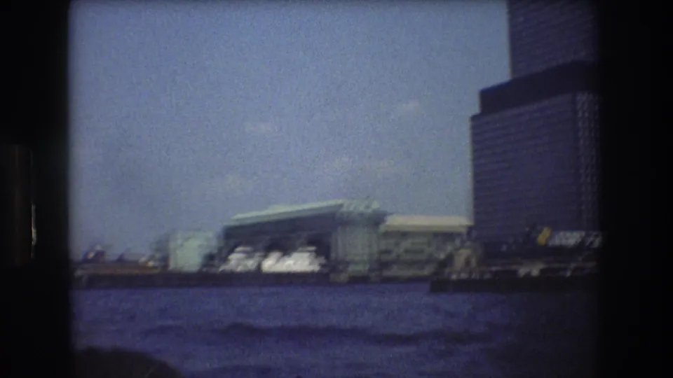 Boat-shot New York City waterfront view showing choppy water, a large terminal structure, a dark tower, and slight blur from handheld 8mm filming.
