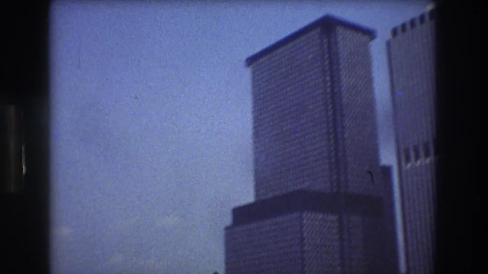 1976 New York City urban waterfront footage frames a tall modern tower and neighboring building against blue sky as the boat viewpoint shifts upward.