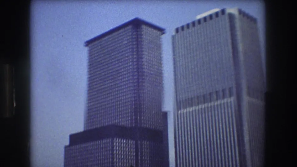 1976 8mm home-movie view of Lower Manhattan skyscrapers from the water, with tall office towers framed by dark film edges.