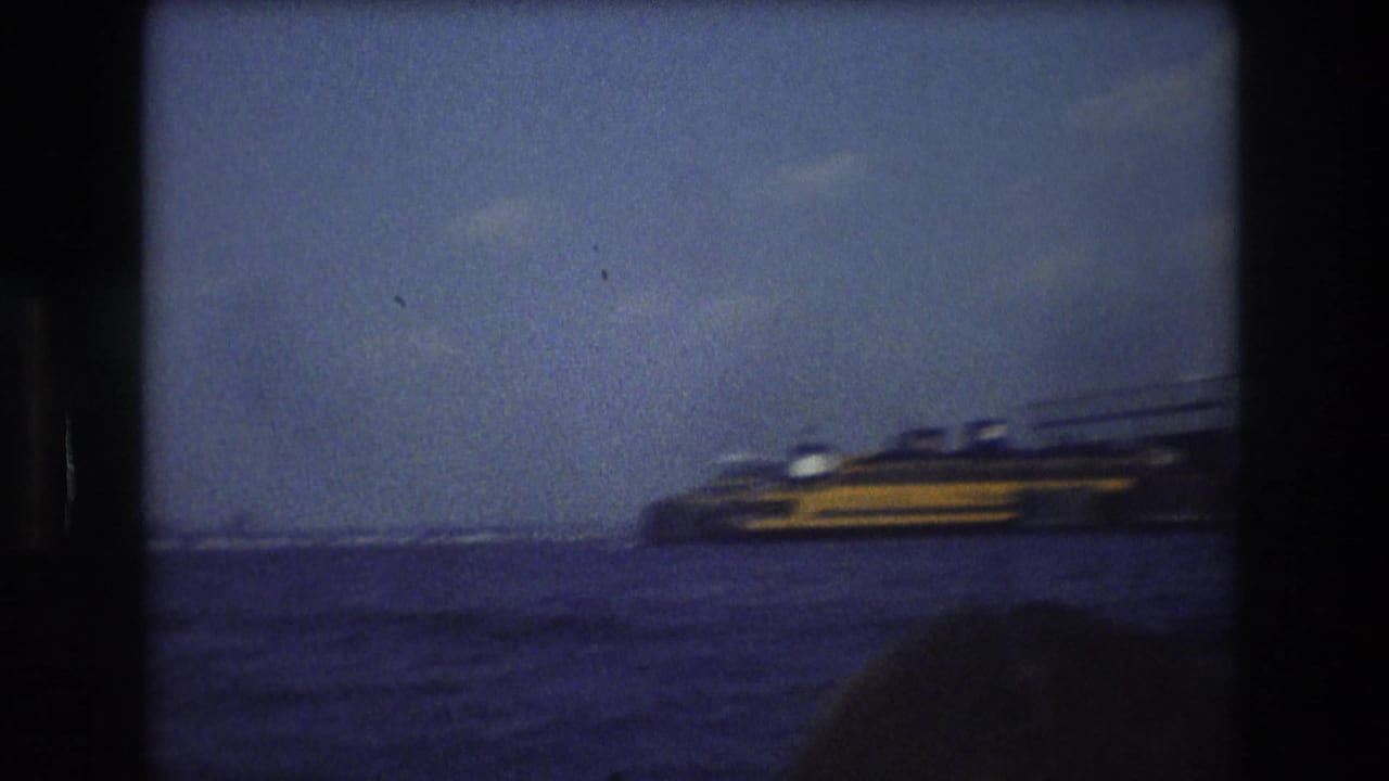 1976 New York City 8mm frame showing a yellow and dark ferry-like vessel on harbor water, with distant skyline forms and the World Trade Center faintly visible through haze.