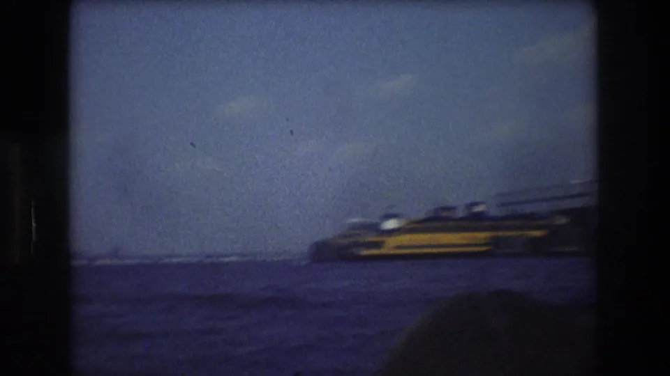 1976 New York City 8mm frame showing a yellow and dark ferry-like vessel on harbor water, with distant skyline forms and the World Trade Center faintly visible through haze.