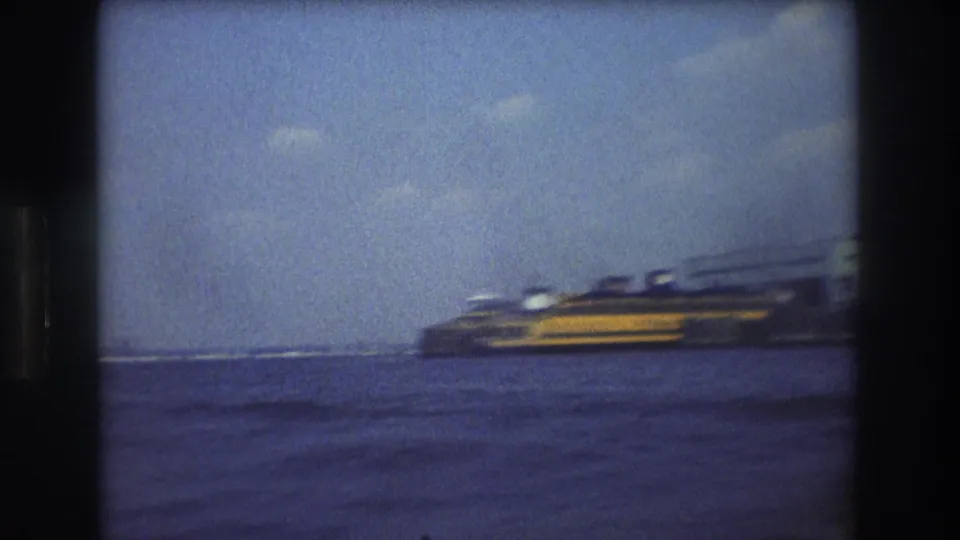 1976 New York City home-movie frame from a boat, showing a yellow and dark vessel moving across harbor water with faint distant waterfront structures in the background.
