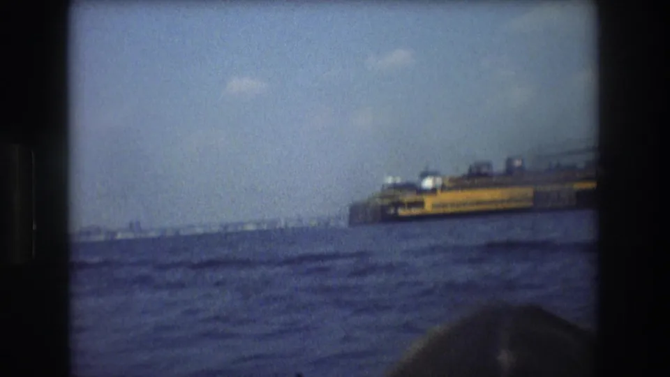 1976 New York City 8mm frame showing a ferry-like vessel across choppy harbor water, with the top of a passenger's head visible at the lower edge.