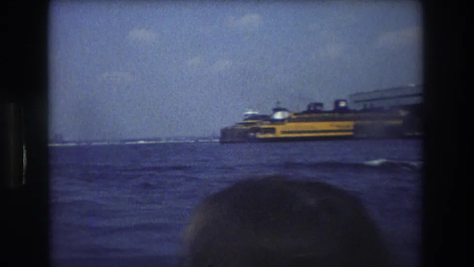 1976 New York City boat-view frame showing a yellow and dark vessel on the harbor, distant waterfront structures, and a partially visible passenger head in the foreground.
