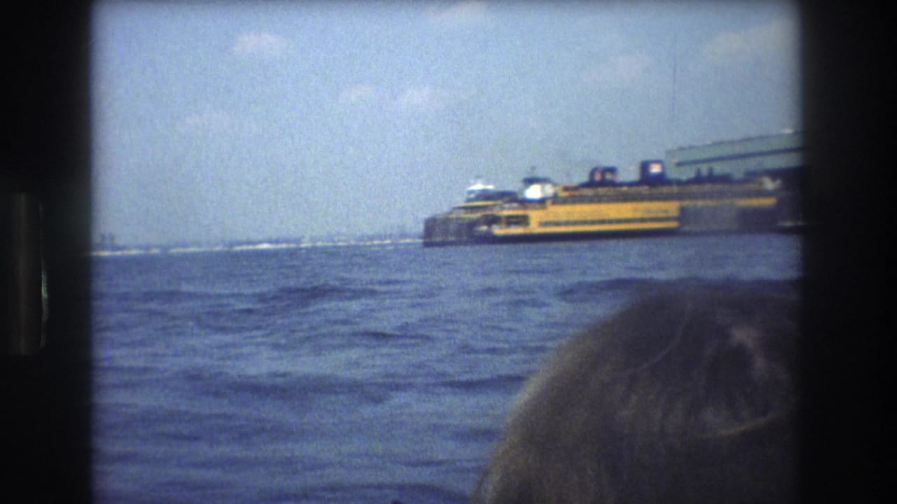 1976 New York City home-movie frame showing a yellow Staten Island Ferry across choppy harbor water, with a dark 8mm border and a partial passenger head near the camera.