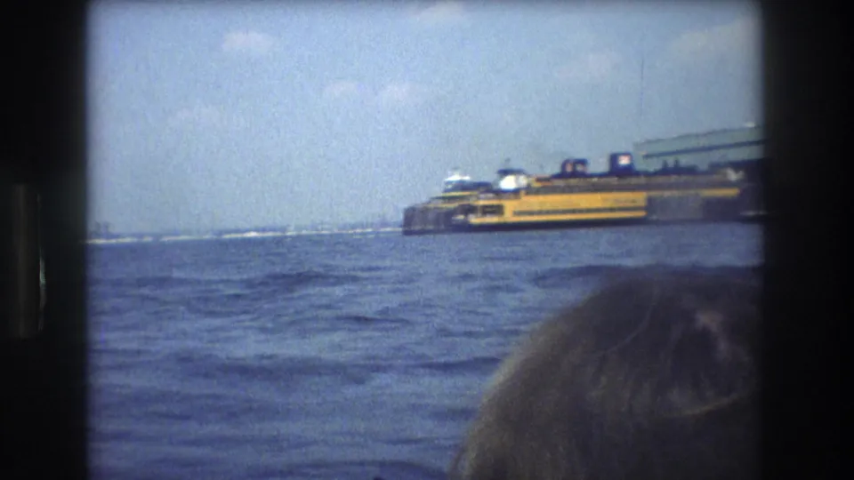 1976 New York City home-movie frame showing a yellow Staten Island Ferry across choppy harbor water, with a dark 8mm border and a partial passenger head near the camera.