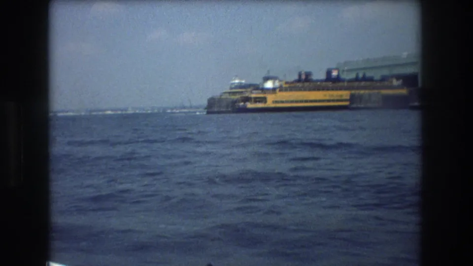 Soft 8mm frame from a moving boat showing the yellow Staten Island Ferry near a low shoreline in New York City, with choppy blue water filling the foreground.