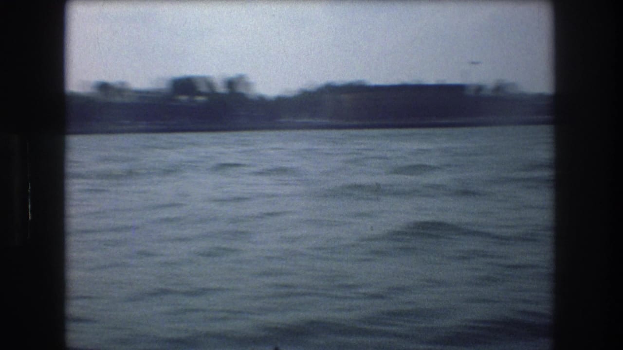 1976 New York City-area 8mm frame from a moving boat, showing rippled water, a low distant waterfront, and dark film borders.