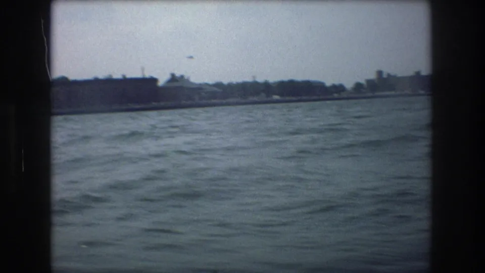 A handheld 8mm boat view across blue-gray water toward a distant New York City-area shoreline with low structures and trees.