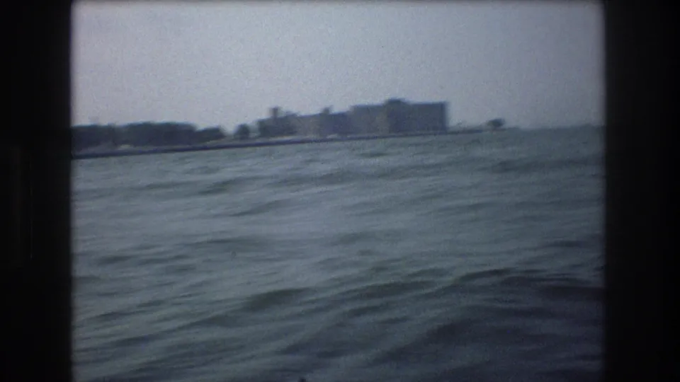 1970s 8mm home-movie frame showing choppy water in the foreground and a hazy distant waterfront structure from a boat perspective.