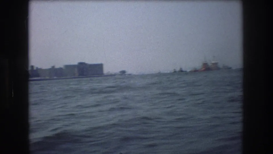 Final 1976 New York City-area boat-view frame with rippled water, low waterfront buildings, and a small distant boat near the horizon.