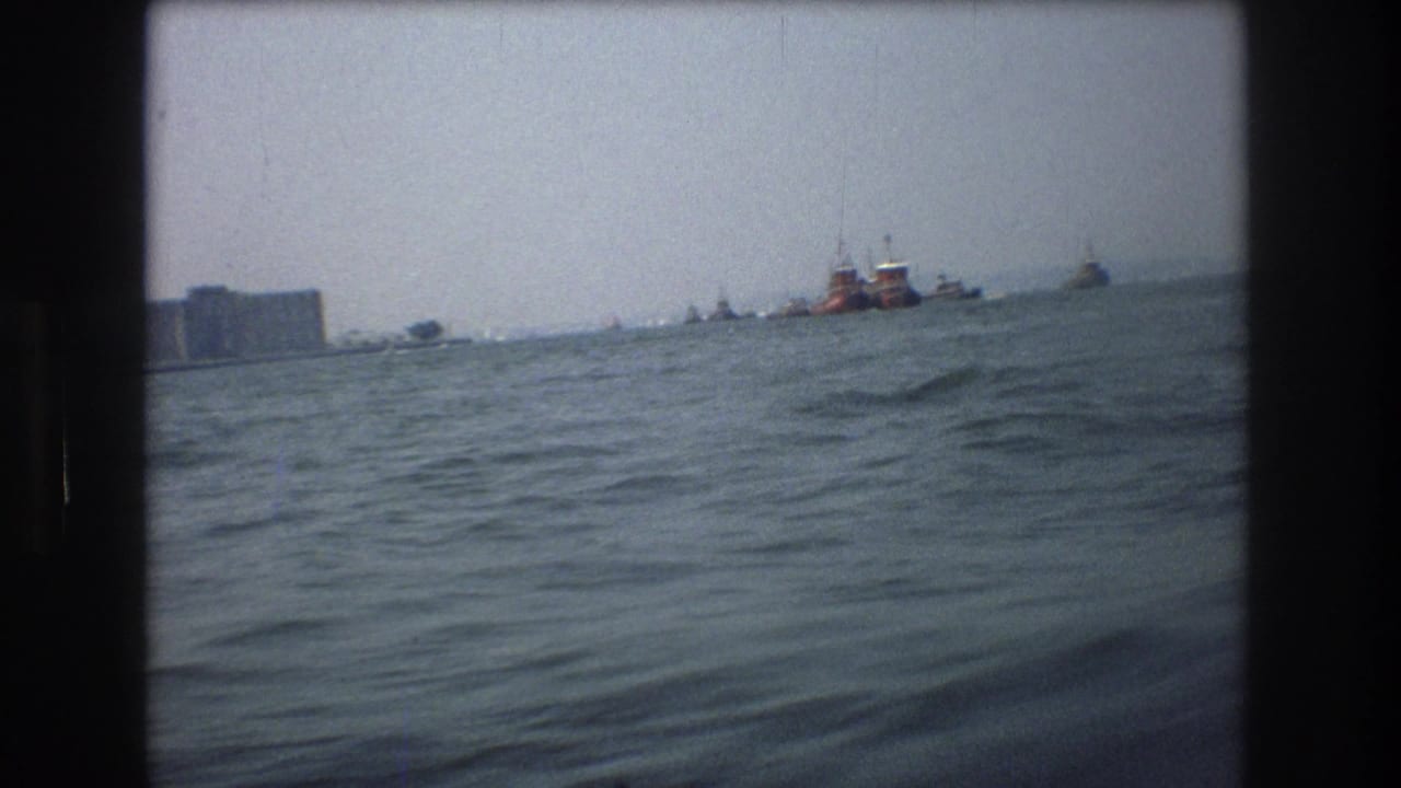 1976 8mm view from a boat in New York City waters, showing rough water, distant vessels, and a hazy shoreline under muted light.