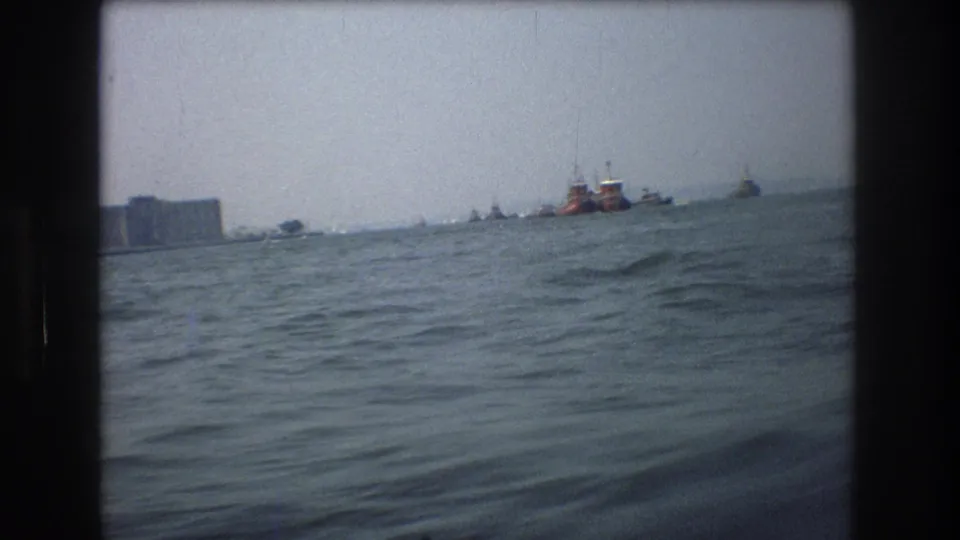 1976 8mm view from a boat in New York City waters, showing rough water, distant vessels, and a hazy shoreline under muted light.