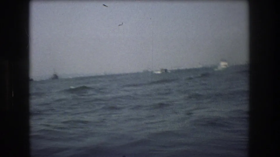 Soft, grainy 8mm frame looking across rough New York City harbor water toward a distant shoreline with indistinct boats and structures.