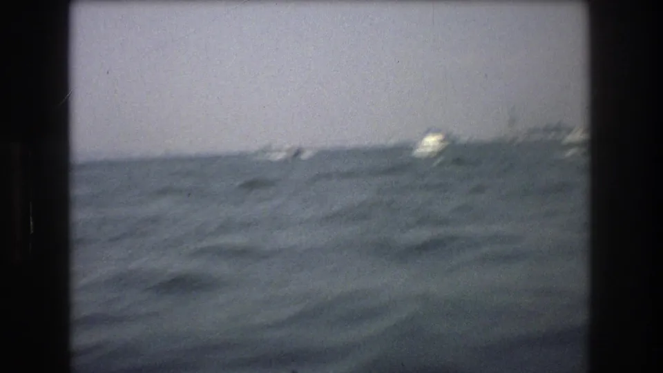 End frame of the 1976 8mm boat view, with rough gray-blue water in the foreground and distant boats or shoreline structures softened by motion and haze.
